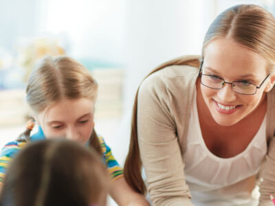 Childcare educator engaging with a small group of young children in a bright early learning centre, sitting together on the floor during a structured activity. The space features child-sized furniture, educational resources and a welcoming indoor environment, reflecting quality early childhood education and care in Victoria.