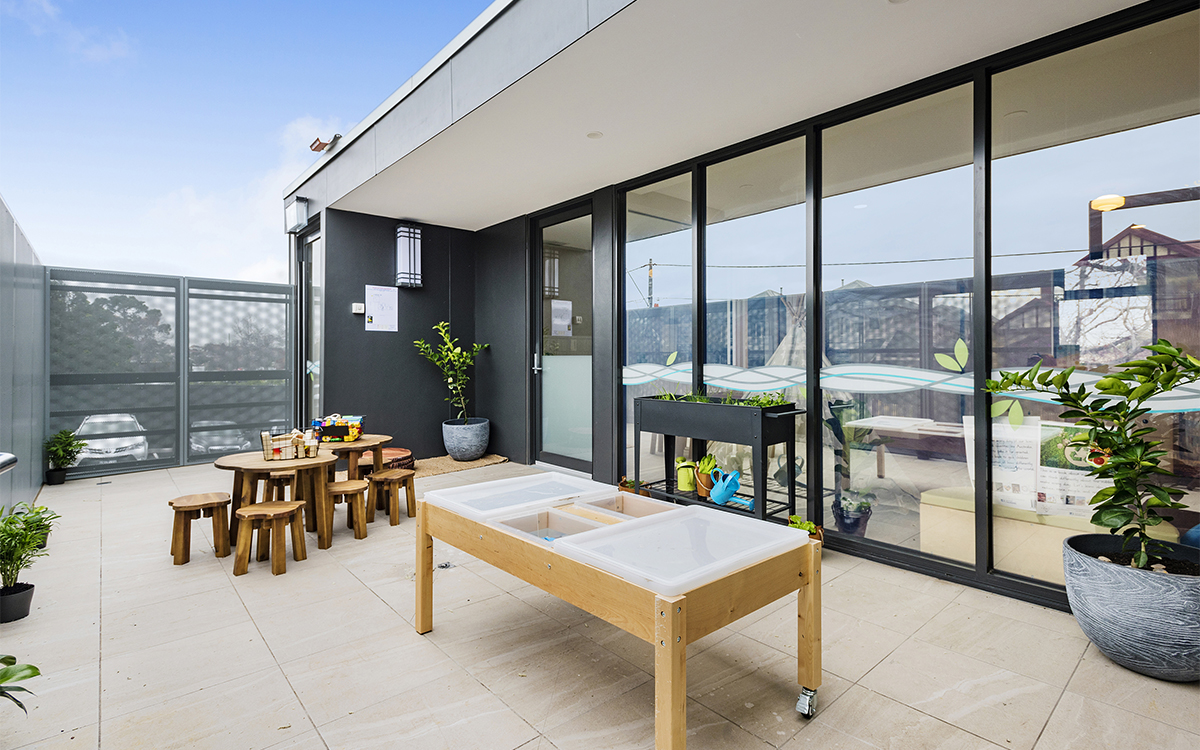 Outdoor learning terrace with children's tables, activity station, planter boxes and glazed building façade.