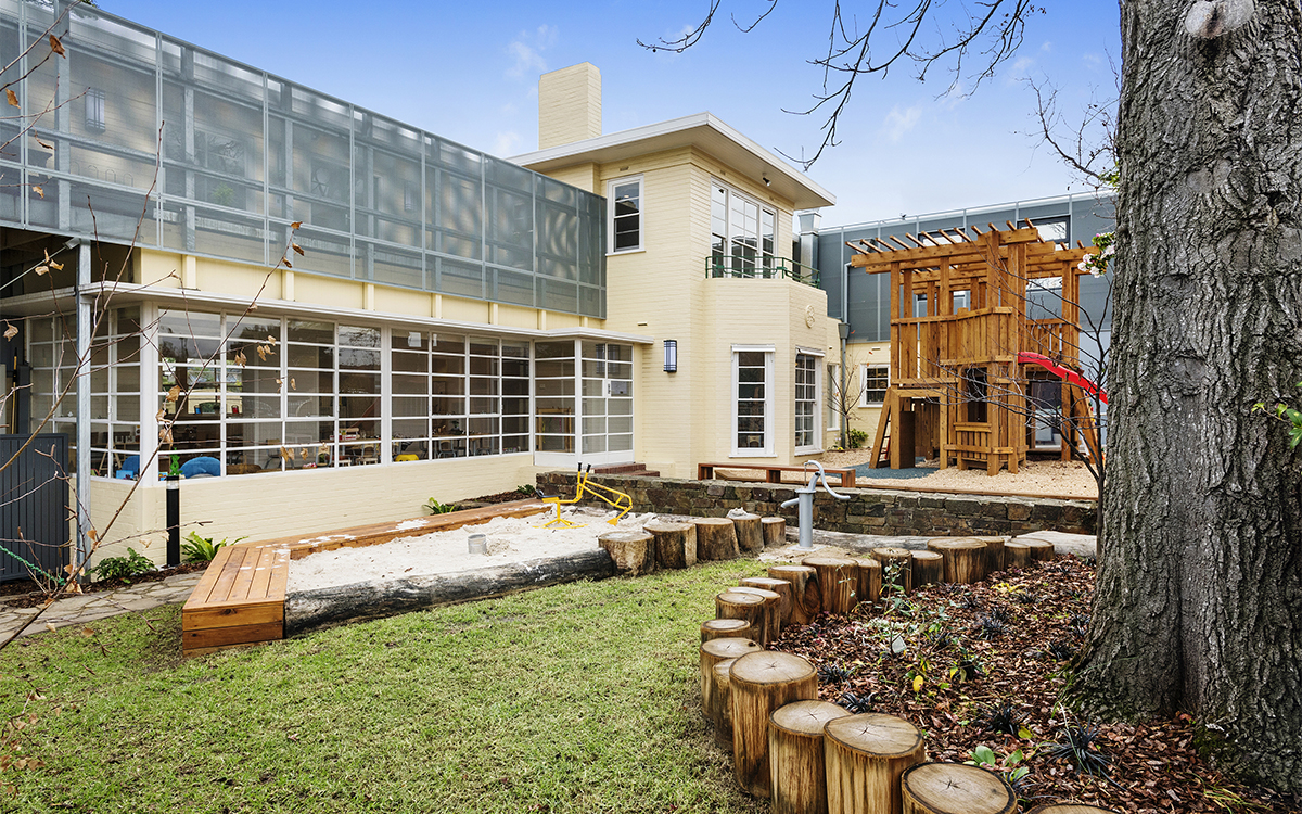 Outdoor play area with lawn, sandpit, play structure and surrounding childcare buildings.