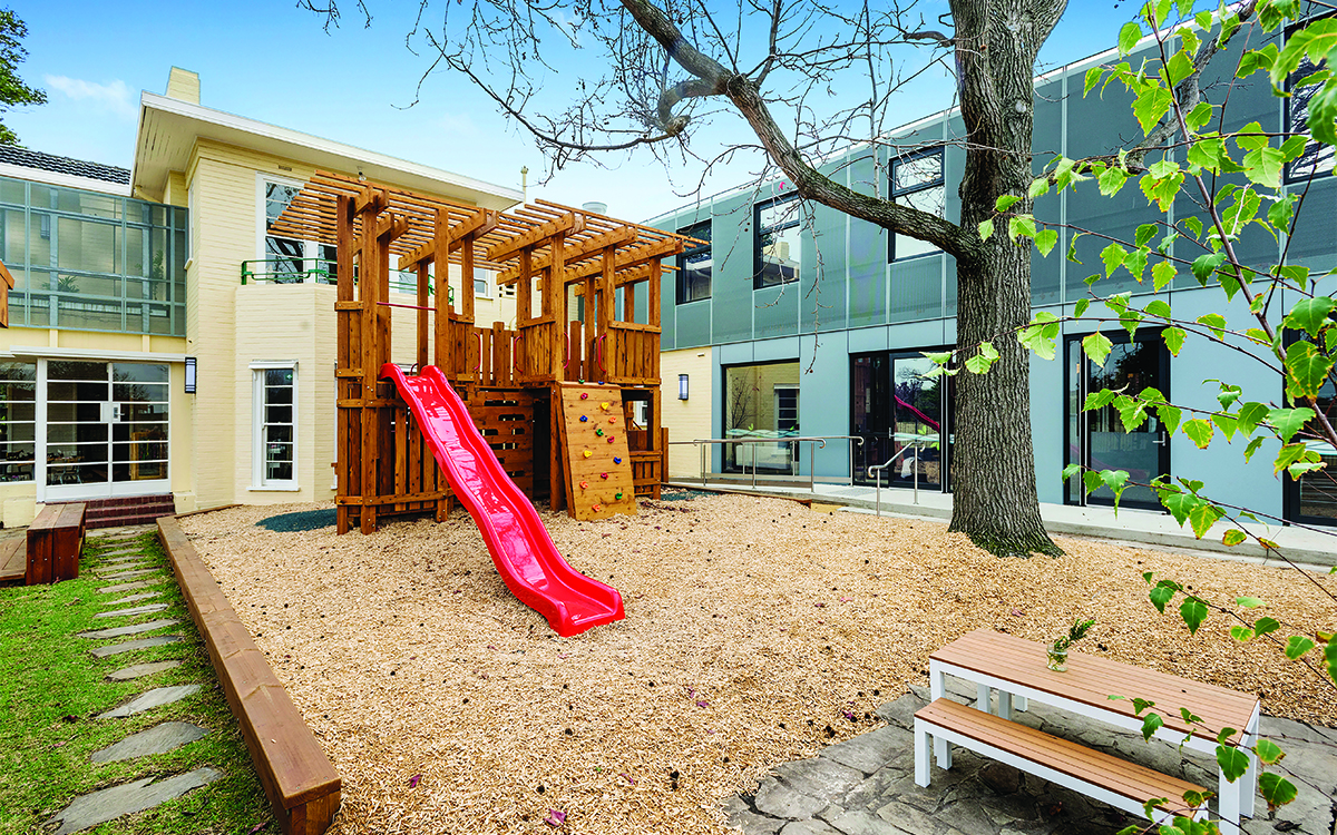 Outdoor playground with climbing structure, slide, seating and mature tree.