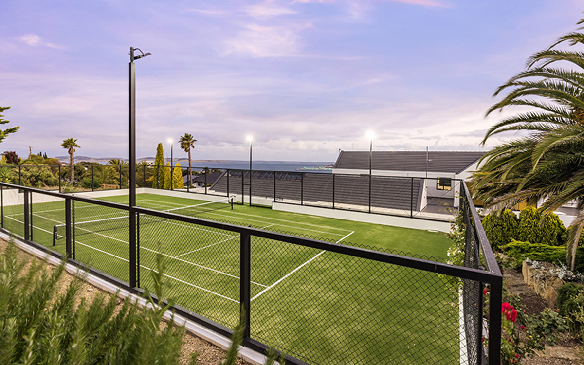 Floodlit full-size tennis court overlooking the coastline and surrounding gardens at dusk.