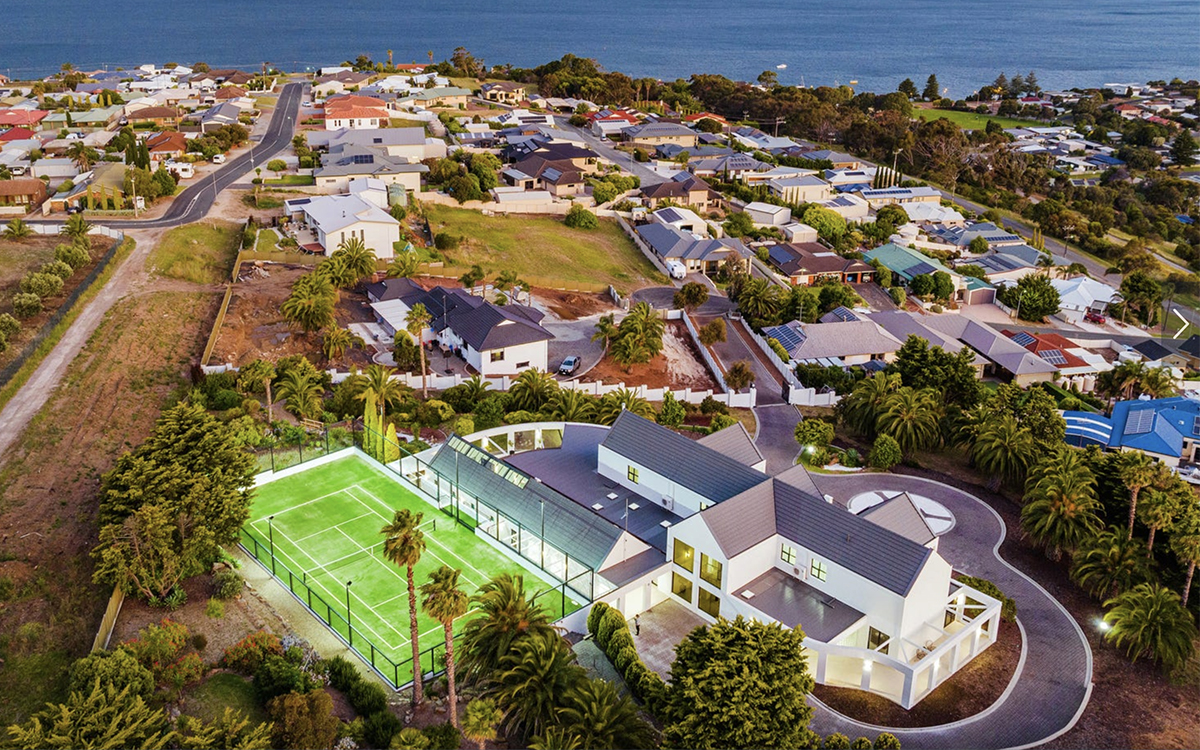 Grand coastal estate with tennis court and landscaped grounds captured from an aerial perspective.