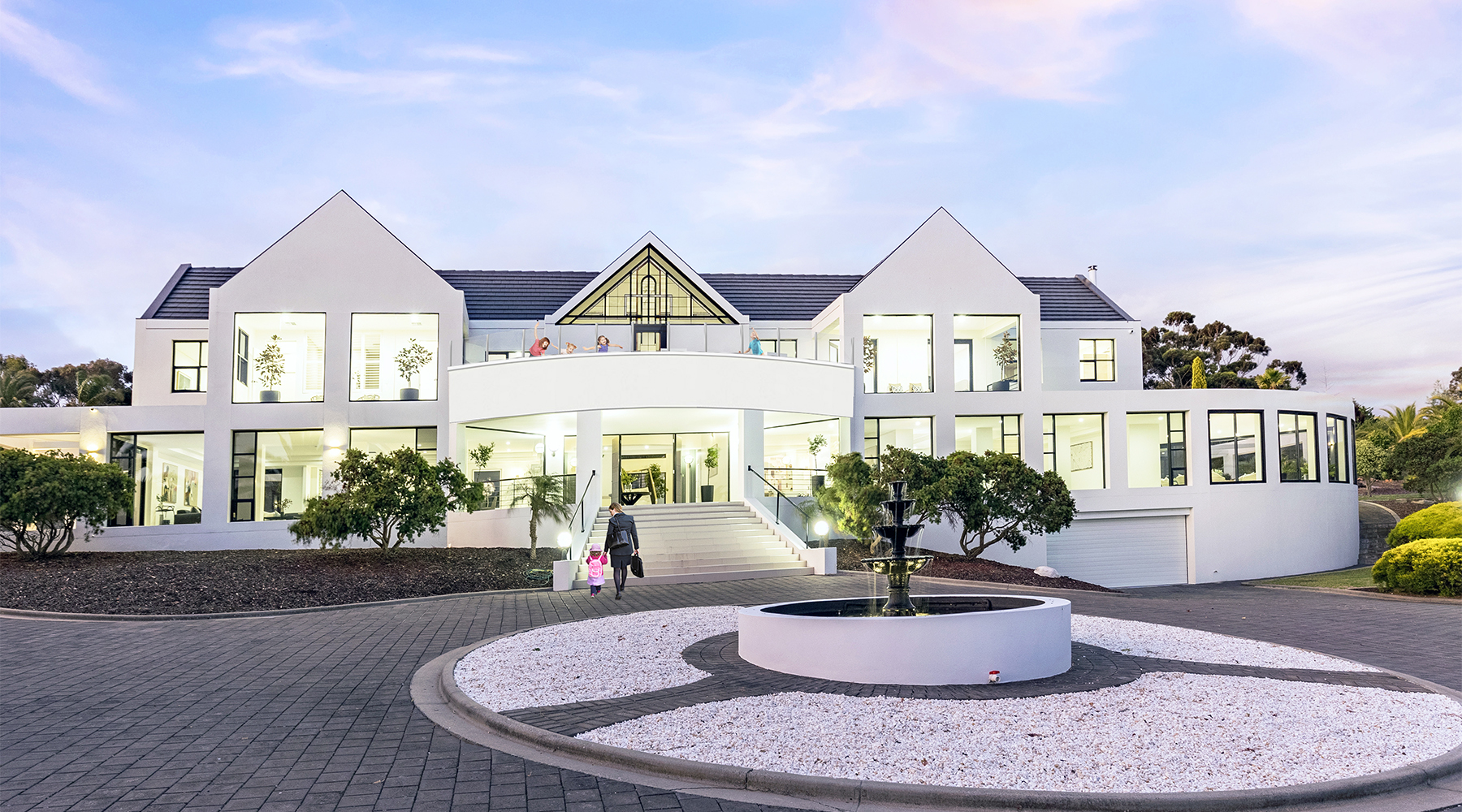 Grand front elevation of the luxury residence with circular driveway, fountain and illuminated façade at dusk.
